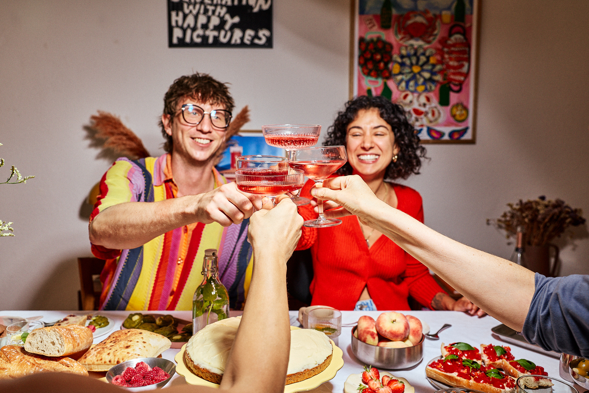 Group of people toasting with pink sparkling wine at a table set with bread, olives, a cake, peaches and tomato bruschetta