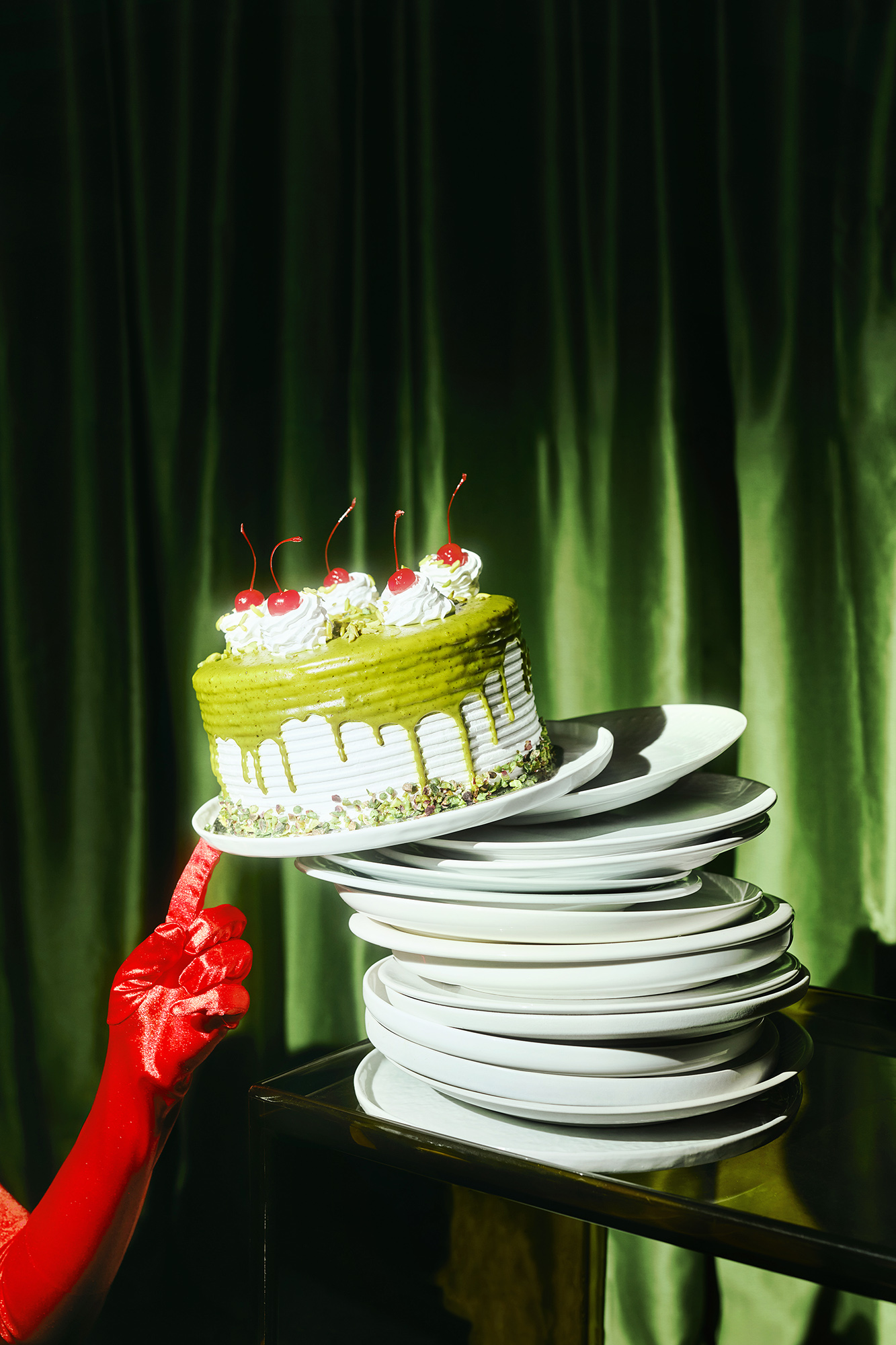 Round matcha cake with a white frosting drip, pistachios and cherries, held above a stack of plates against a green curtain