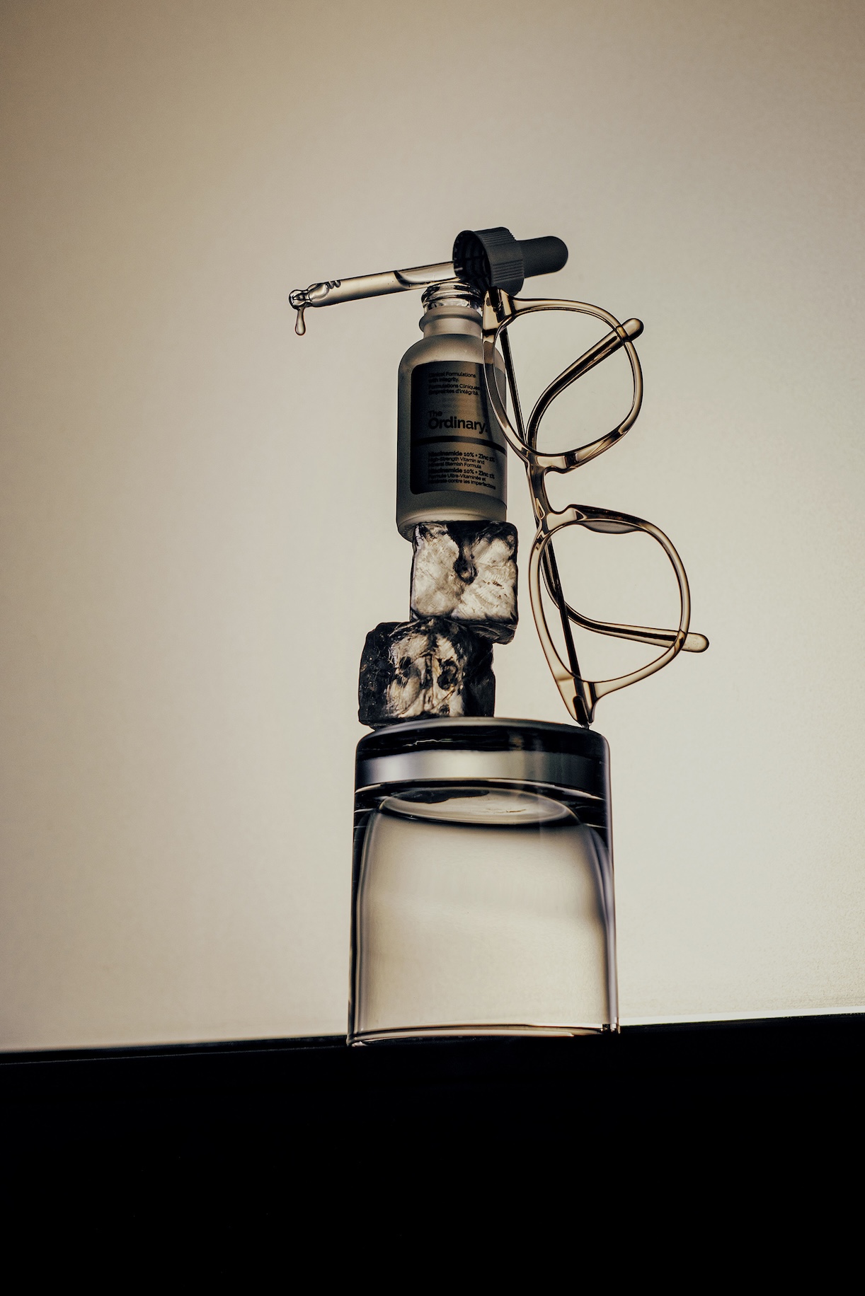 Still life of a dropper bottle, a pair of glasses and ice cubes balanced on top of a glass of water