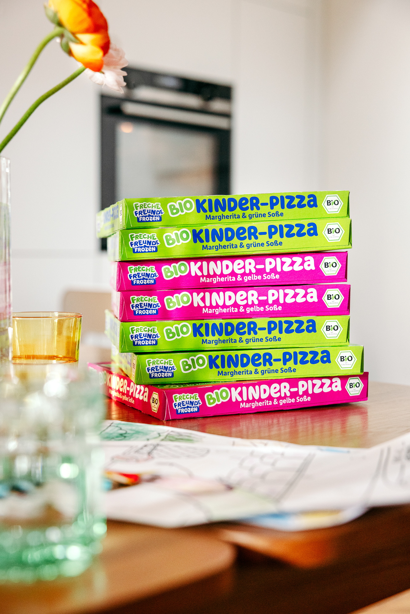 Stacked boxes of Bio Kinder-Pizza in green and pink packaging on a kitchen table with a vase of flowers