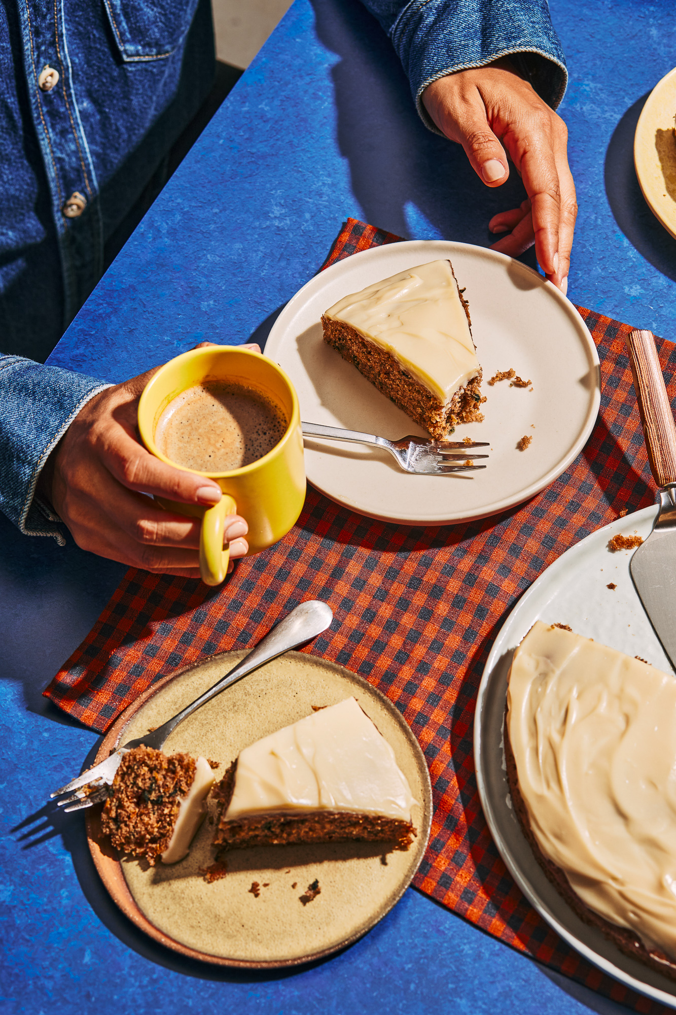 Overhead view of two hands eating carrot cake with frosting, with a yellow coffee mug on a checkered table runner