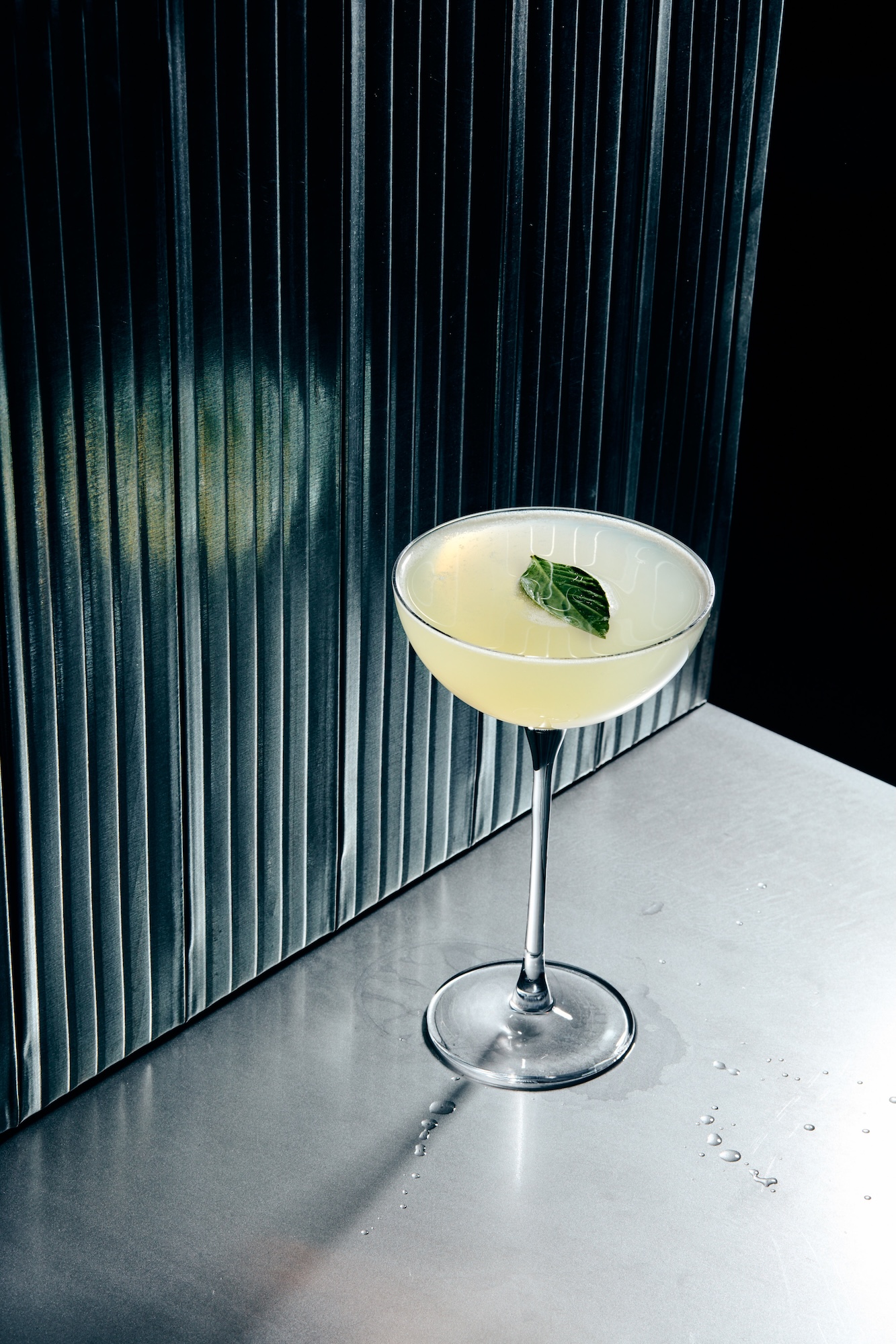 Cocktail glass with a pale yellow drink garnished with a mint leaf on a metal bar counter