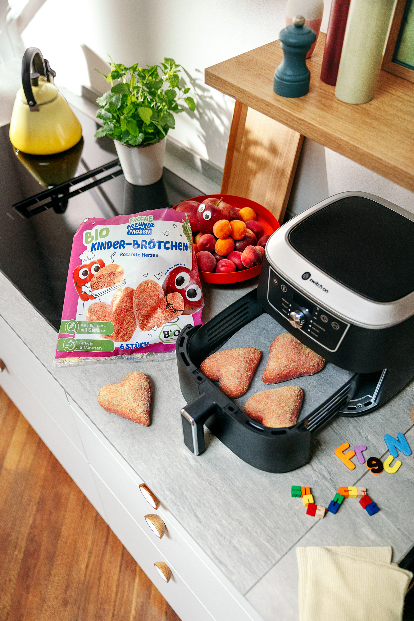 Kitchen counter with an air fryer full of heart-shaped rolls, next to a fruit bowl and a kettle