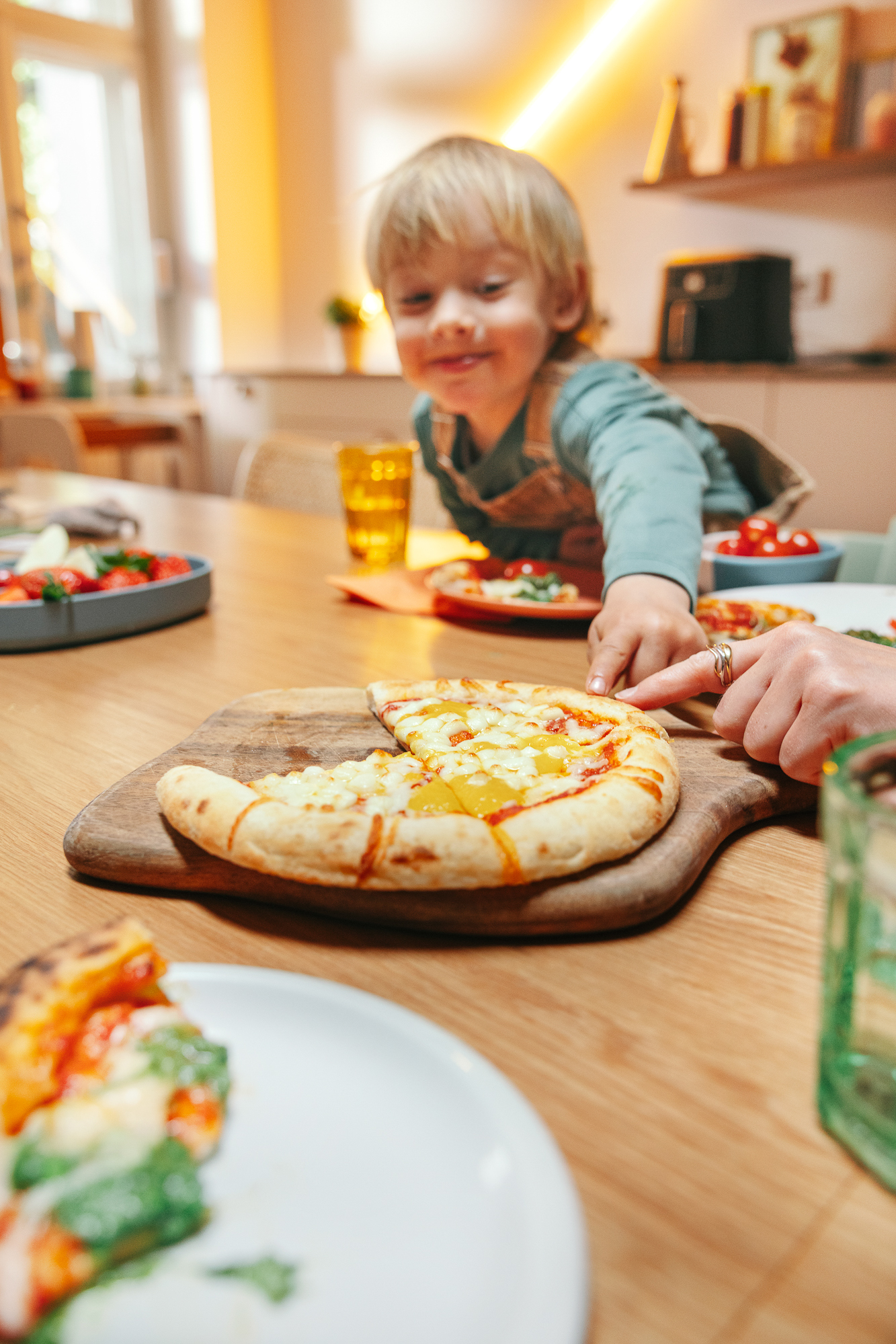 Toddler smiling at the table next to a freshly baked pizza on a wooden board, a hand pointing at it