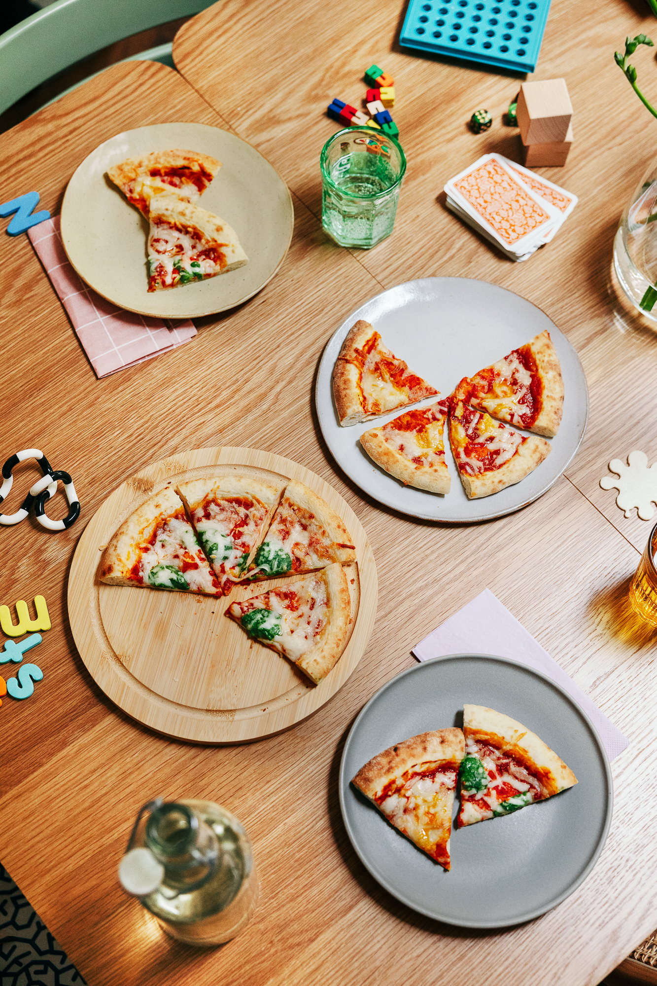 Four plates of sliced pizza on a wooden table, surrounded by toys and playing cards