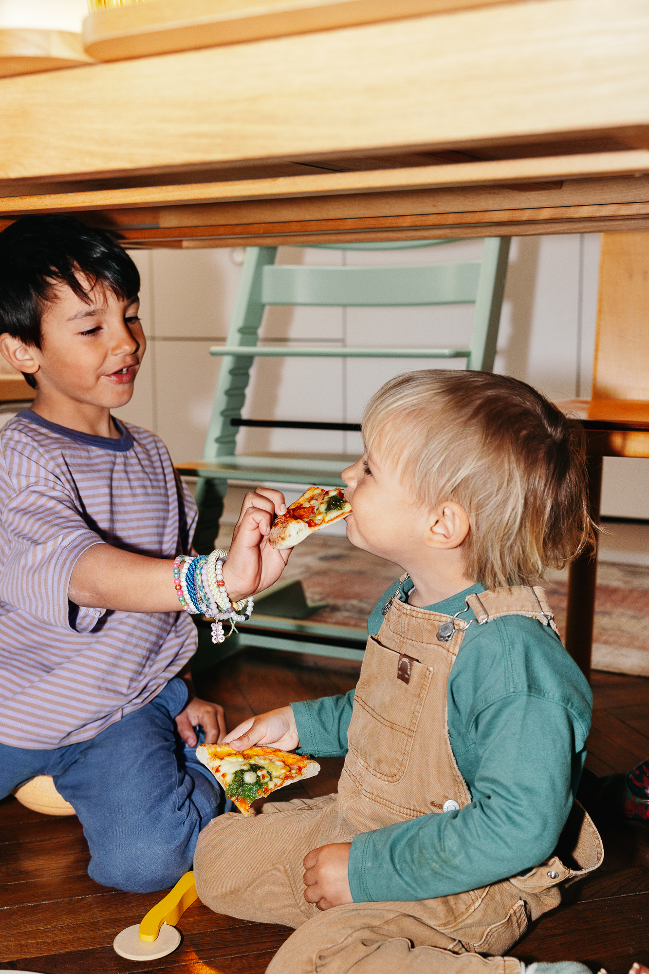 Two children sitting under a table, an older child feeding a younger one a slice of pizza