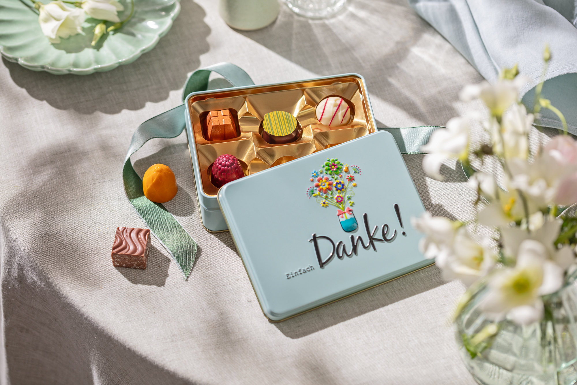Open box of chocolates reading Einfach Danke, surrounded by loose chocolates, flowers and a green ribbon